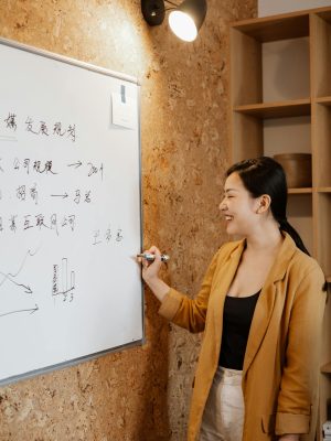 A smiling Asian woman presents business strategies on a whiteboard in an office setting.