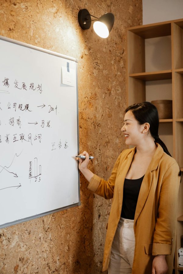 A smiling Asian woman presents business strategies on a whiteboard in an office setting.