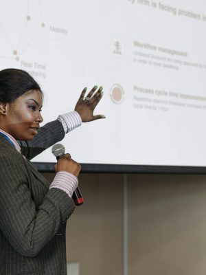 Businesswoman presenting with microphone, discussing improvements with a slide projection in a conference room.